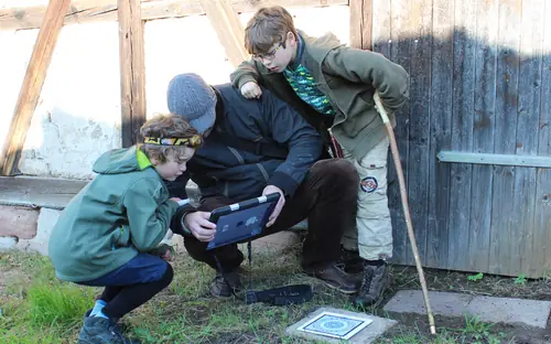 Kinder und ein Erwachsener lösen beim Outdoor-Exitgame „Das magische Portal“ in Oberkirch gemeinsam eine Aufgabe am Tablet an einer alten Fachwerkscheune